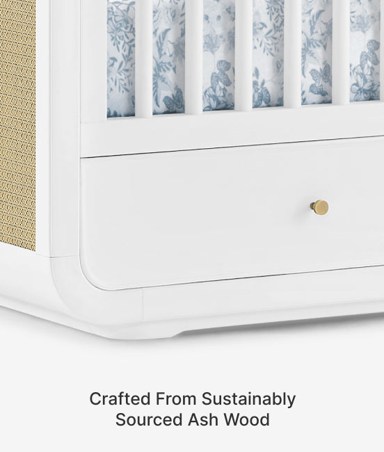 White crib with blue floral patterned bedding on a white background, featuring text 'Crafted From Sustainably Sourced Ash Wood'.