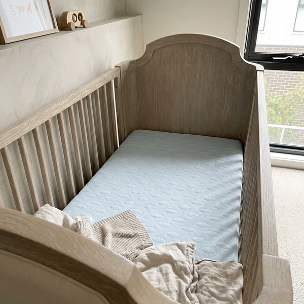 Wooden crib with a light blue mattress and beige blanket in a room with a window.