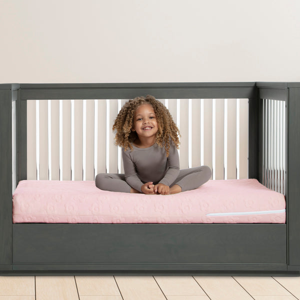 Child sitting on a pink mattress in a gray crib with a plant in the corner.