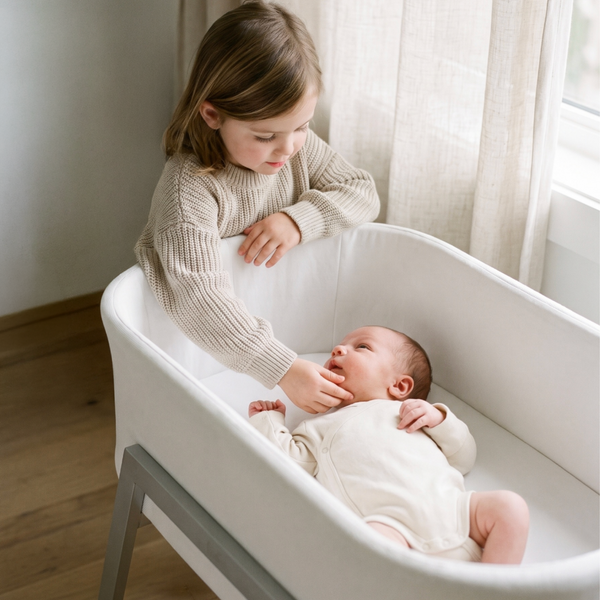 A newborn baby in a cream onesie lies in a white bassinet with gray legs, while a young girl in a matching cream ribbed sweater leans over the side of the bassinet in a softly lit room with sheer curtains and wood flooring.