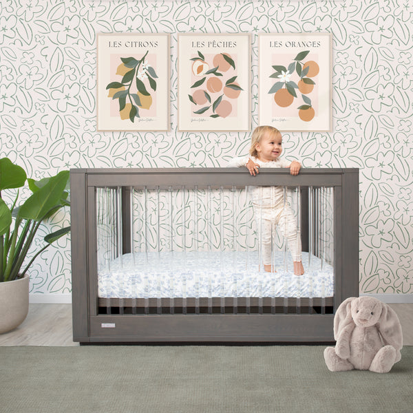 Child peeking over a crib with decorative wall art and a plush toy on a patterned wall.