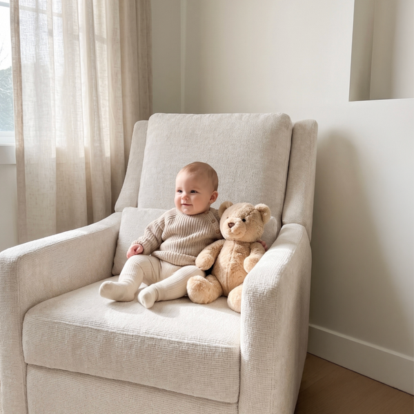A baby in a beige knit outfit sits in a cream upholstered armchair next to a teddy bear, in a softly lit room with neutral walls and curtains.