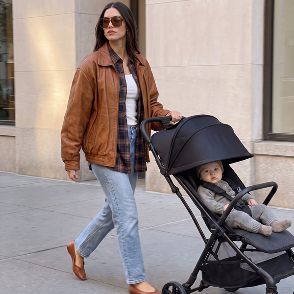 A woman wearing a brown leather jacket, white tank top, and blue jeans pushes a black stroller with a baby inside, walking on a city sidewalk next to a beige building.