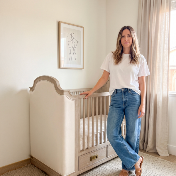 A woman stands in a softly lit nursery beside a light wood crib, wearing a white T-shirt, wide-leg blue jeans, and brown sandals. The room has cream walls, beige curtains, a textured rug, and a framed minimalist line drawing.