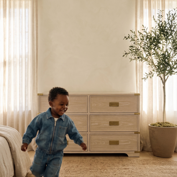 A toddler wearing a blue denim jumpsuit runs happily on a woven rug in a bright, modern bedroom with a light wood dresser, sheer curtains, and a potted olive tree.