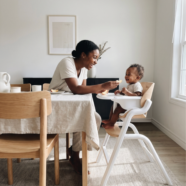 A woman sits at a wooden dining table with a child in a white high chair, sharing a meal in a bright, modern dining room.